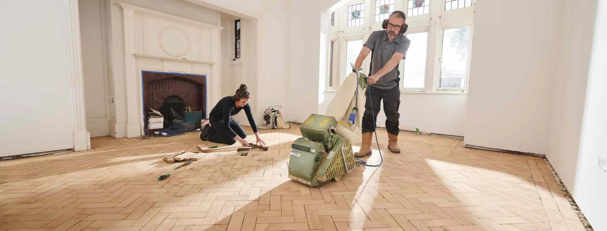 A technician from "Sanding Wood Floors" is using a Lagler HUMMEL&reg; belt sander to sand a wooden floor in a living area in Burnham, UK. The machine has a single-phase AC motor with 2.2 kW power, 230 V voltage, 50 Hz frequency, 200 mm drum width, 2400 rpm drum speed, 200x750 mm sanding belt, and weighs 79 kg. It features a powerful LED lighting system, extremely low dust emissions, quick belt changes, and 3x adjustable pressure.