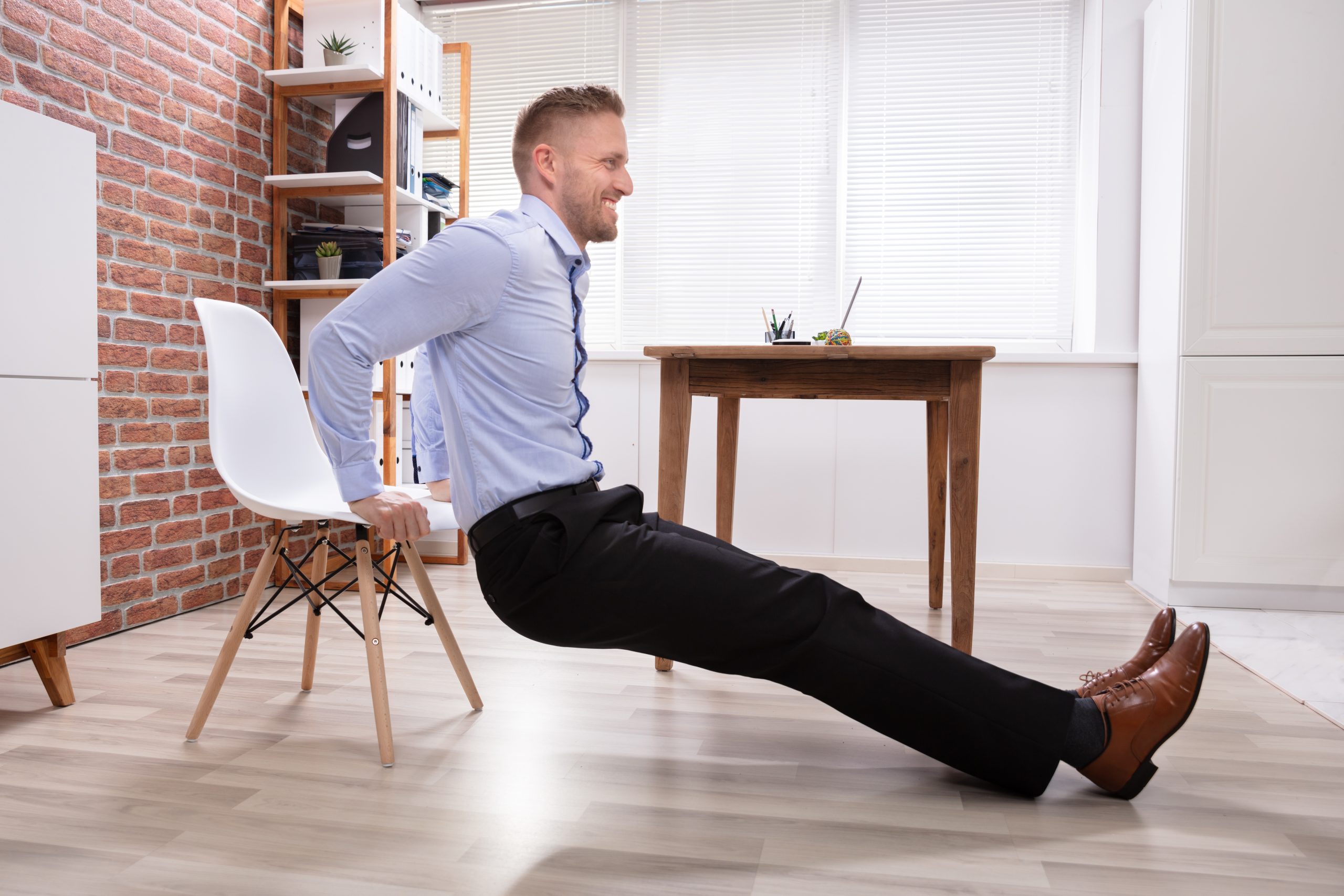 A man in a blue dress shirt and black trousers performs tricep dips using a white chair in a home office with a brick accent wall, wooden shelves, and a desk. The beautifully sanded and refinished wooden floor adds a polished look to the modern workspace, highlighting the expert work done by "Sanding Wood Floors."