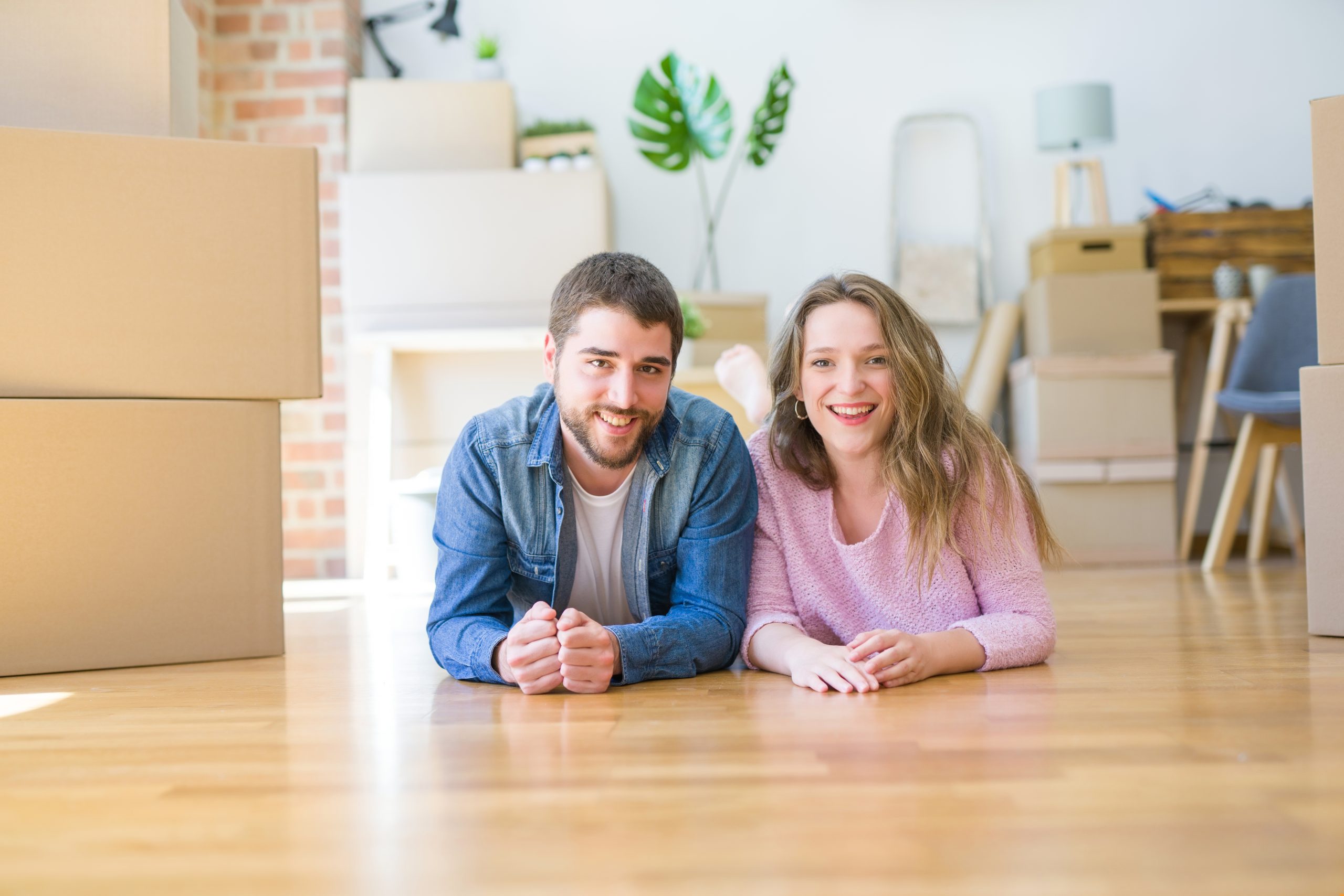 A smiling couple lying on a beautifully refinished wooden floor in their new home, surrounded by moving boxes. The light wood floor shines with a polished finish, showcasing the expert floor sanding and refinishing done by "Sanding Wood Floors."