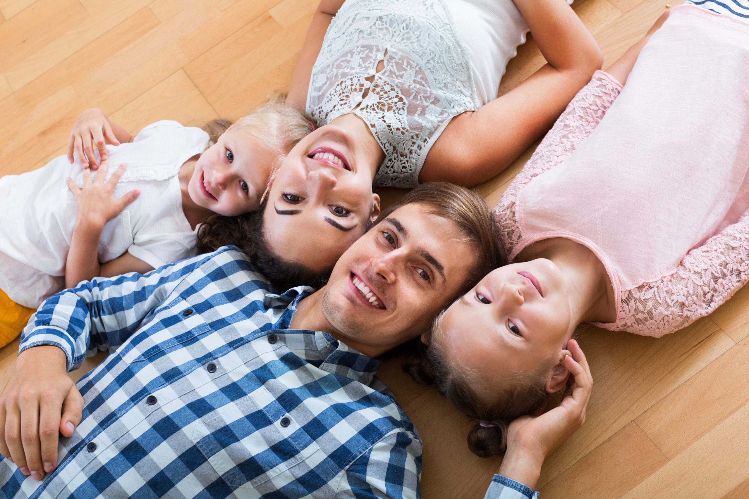 A cheerful family of four lies on their beautifully refinished wooden floor, smiling up at the camera. The parents and two young children look happy and comfortable, showcasing the quality of the floor sanding and refinishing done by "Sanding Wood Floors."