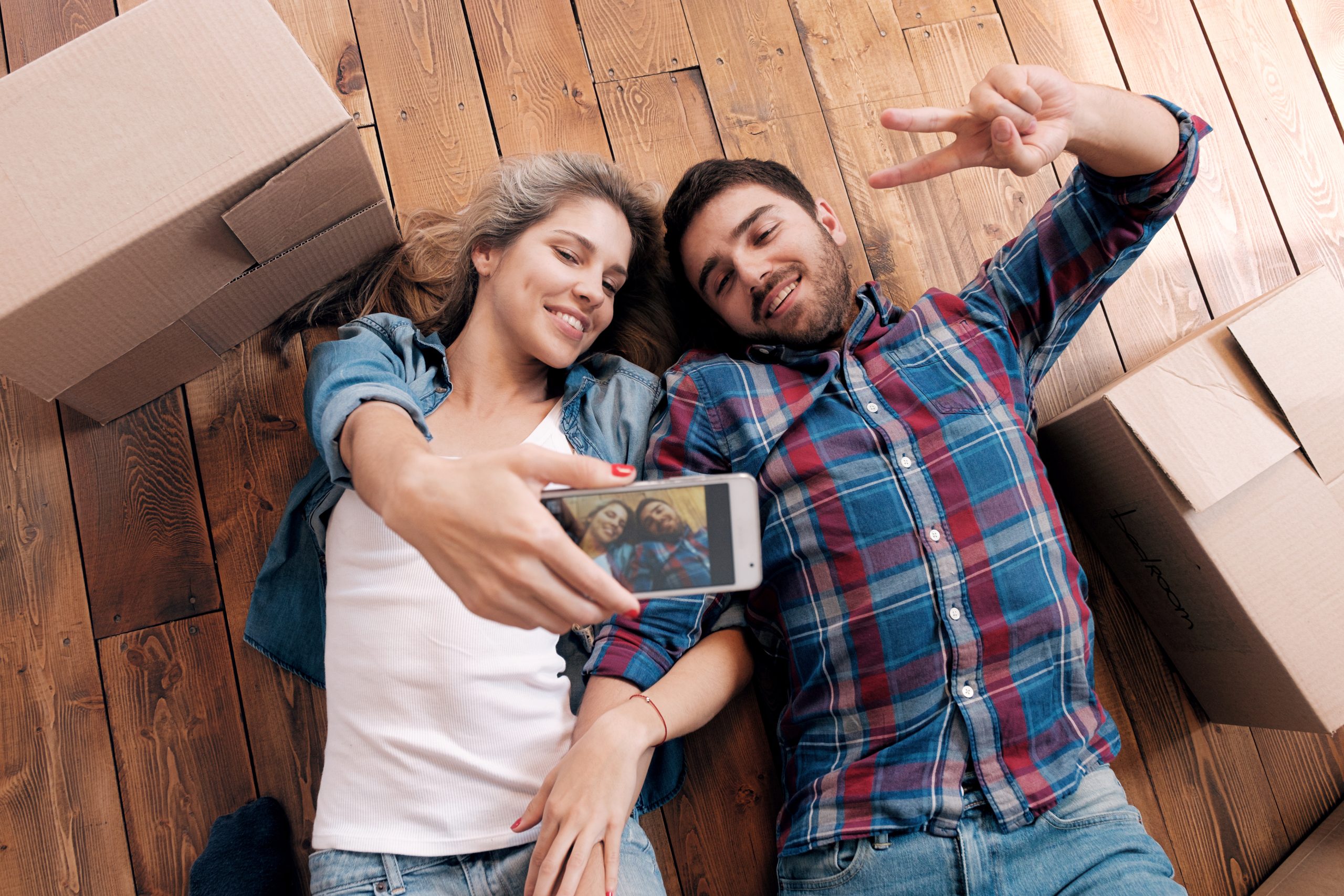 A young couple is lying on their backs on a beautifully refinished wooden floor, surrounded by cardboard boxes. They are smiling and taking a selfie with a smartphone, with the man flashing a peace sign.