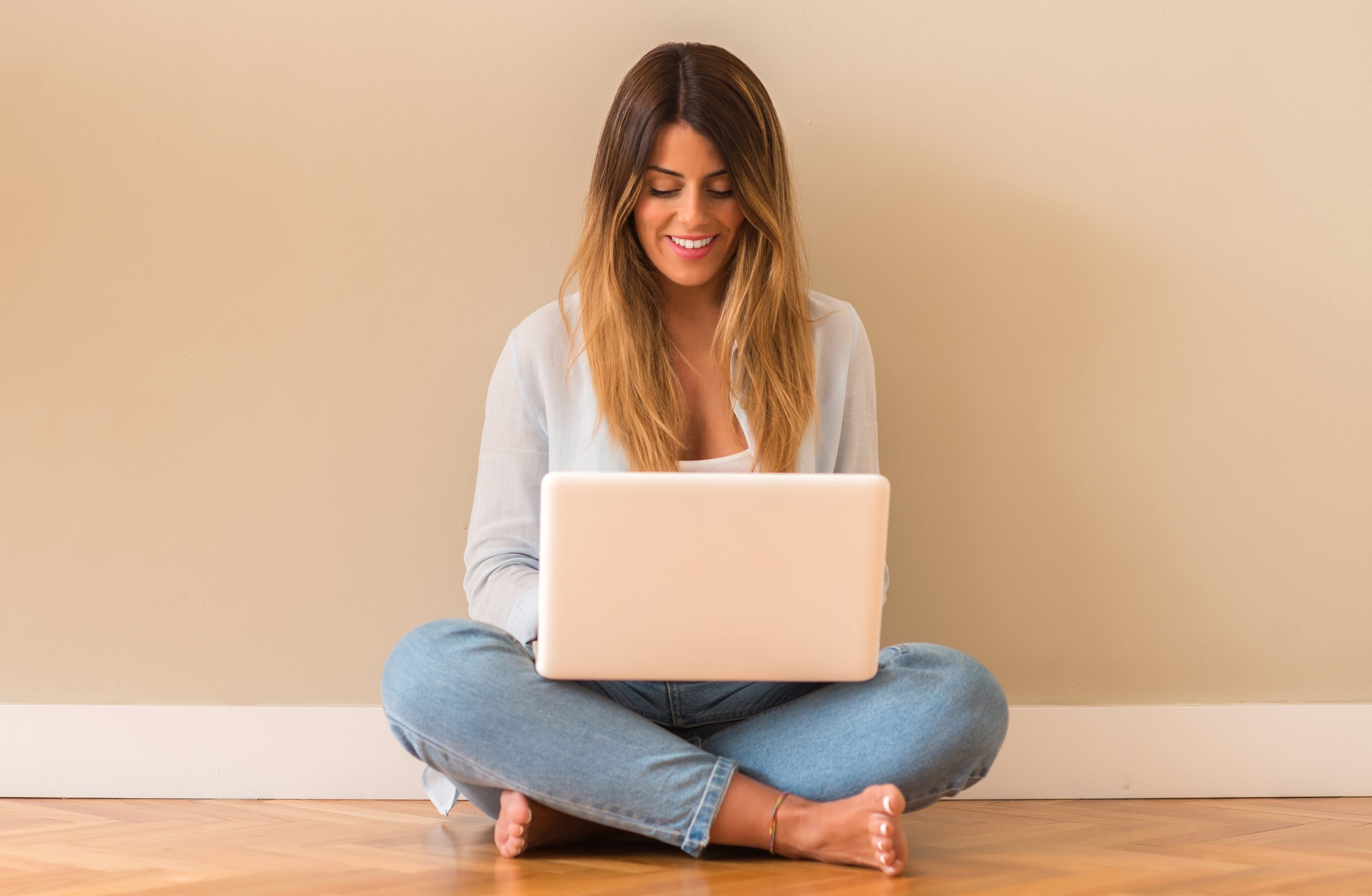 A young woman sitting cross-legged on the floor, smiling as she works on her laptop. The beautifully sanded and refinished wooden floor beneath her, done by "Sanding Wood Floors," adds warmth and elegance to the cozy space.