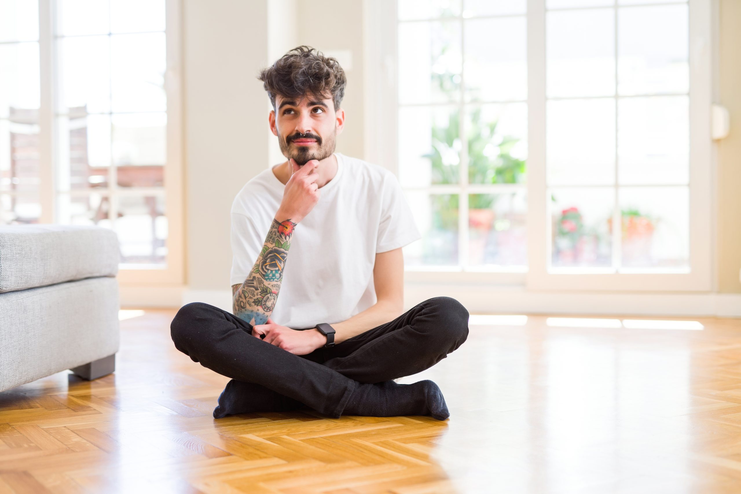 A thoughtful man with a tattooed arm sitting cross-legged on a beautifully refinished wooden parquet floor, contemplating in a bright, sunlit room. The smooth and polished floor showcases the expert floor sanding and refinishing work done by "Sanding Wood Floors."