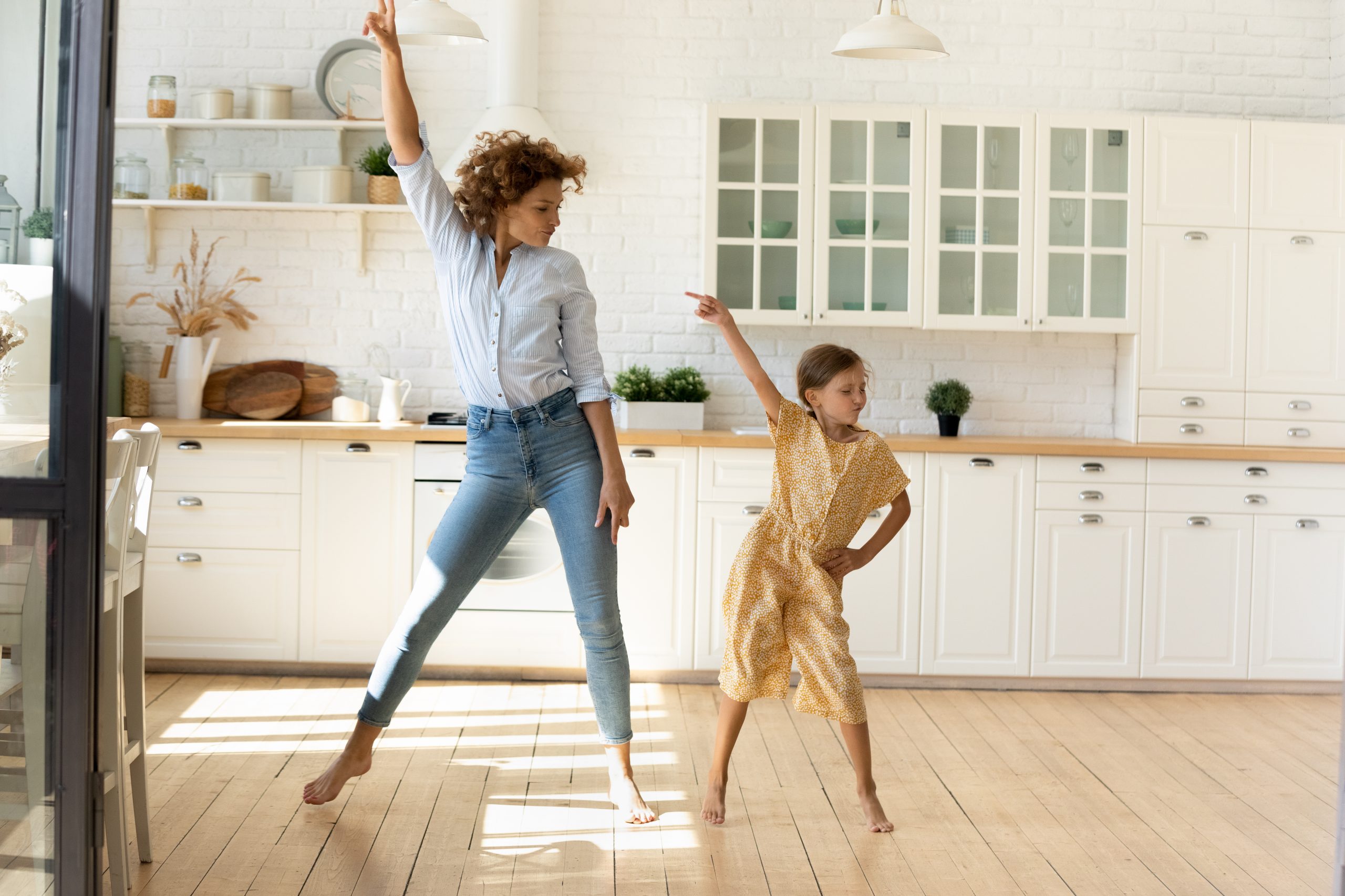 A mother and daughter dance joyfully in their bright kitchen, showcasing the beautifully sanded and refinished wooden floors. The warm tones of the wood complement the light-filled space, reflecting the quality work done by Sanding Wood Floors.