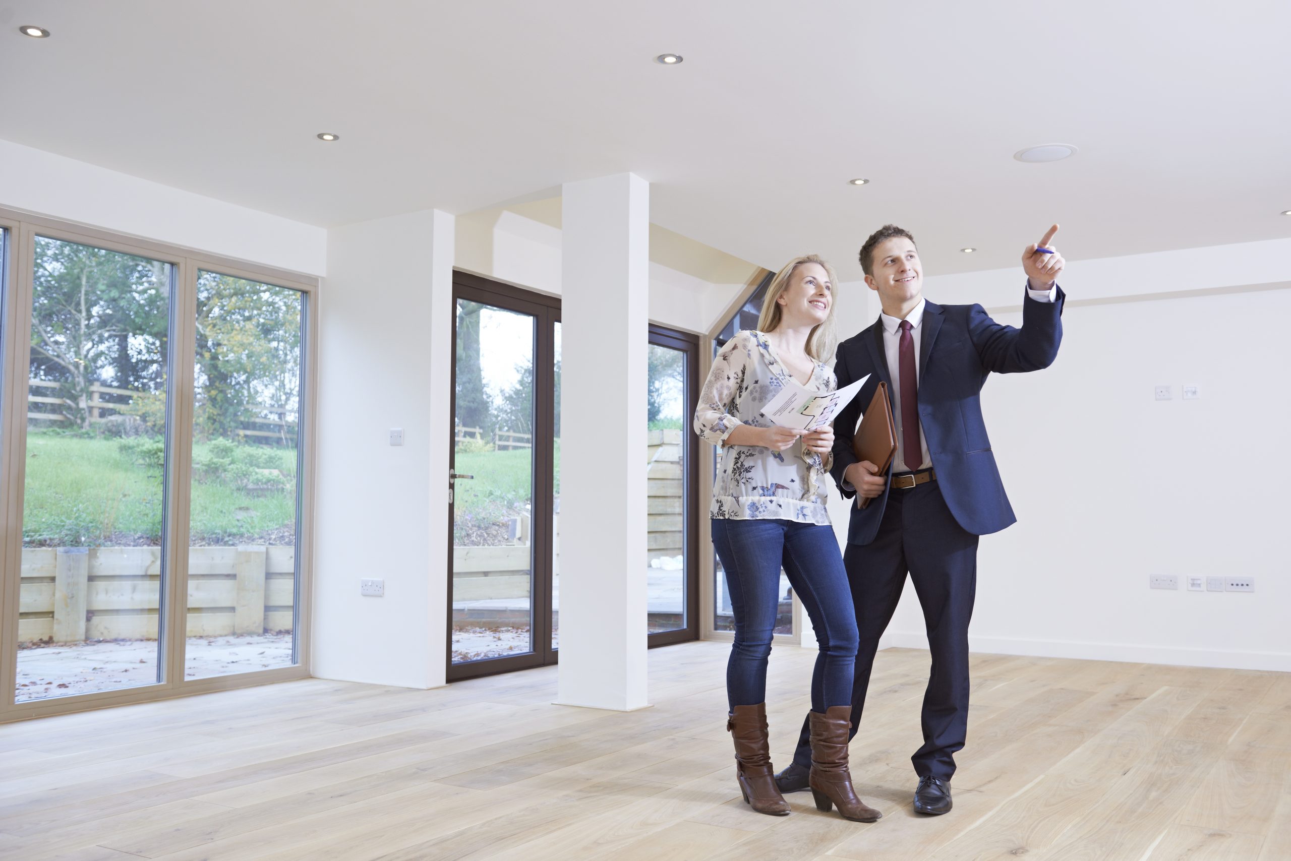 A happy couple being guided by a professional real estate agent in a bright, newly renovated room with large windows and freshly refinished wooden floors. The floors, expertly sanded and refinished by "Sanding Wood Floors," add a warm and inviting atmosphere to the space.