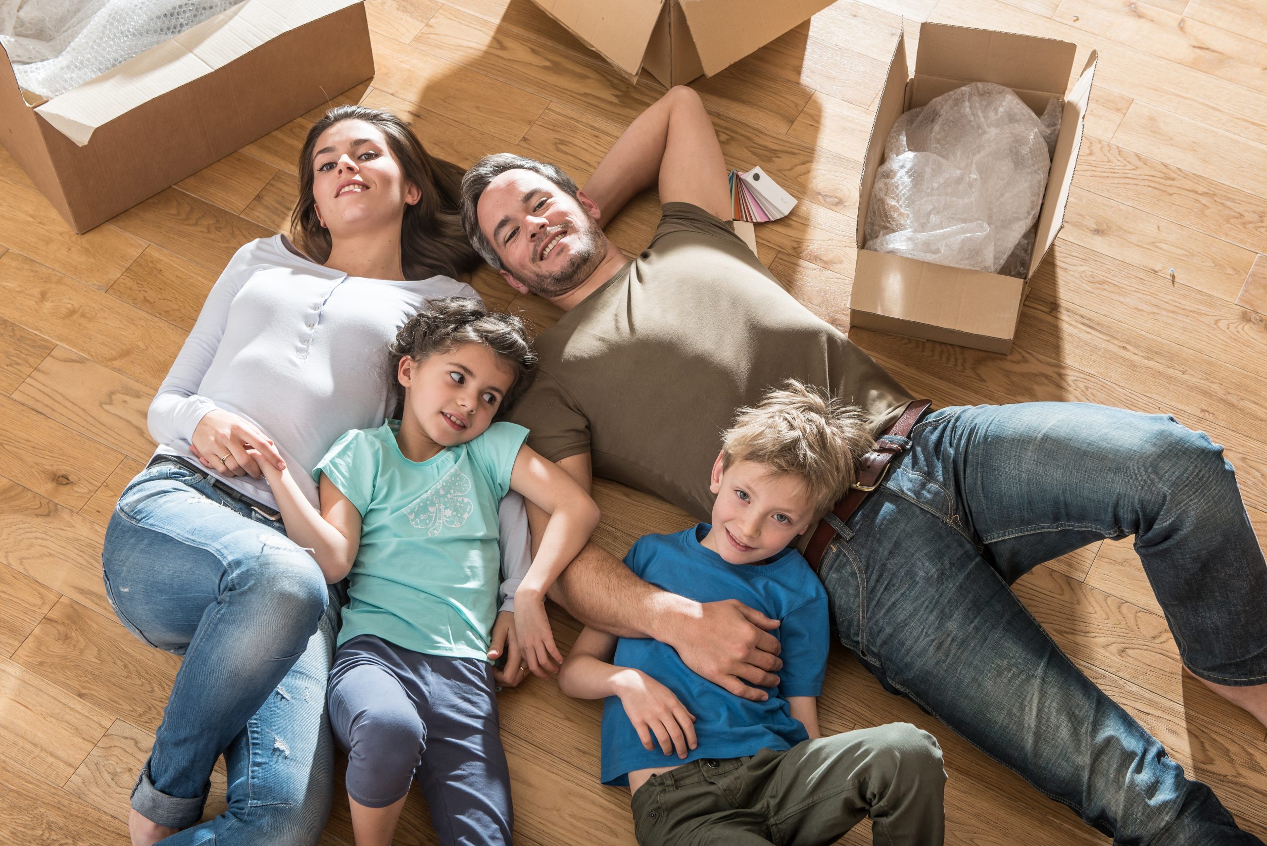 A happy family of four is lying on a beautifully refinished wooden floor, surrounded by unpacked cardboard boxes. The parents and their two children are smiling and enjoying a moment of relaxation together, celebrating their new home. The warm tones of the wooden floor add to the comfort and joy of the scene.