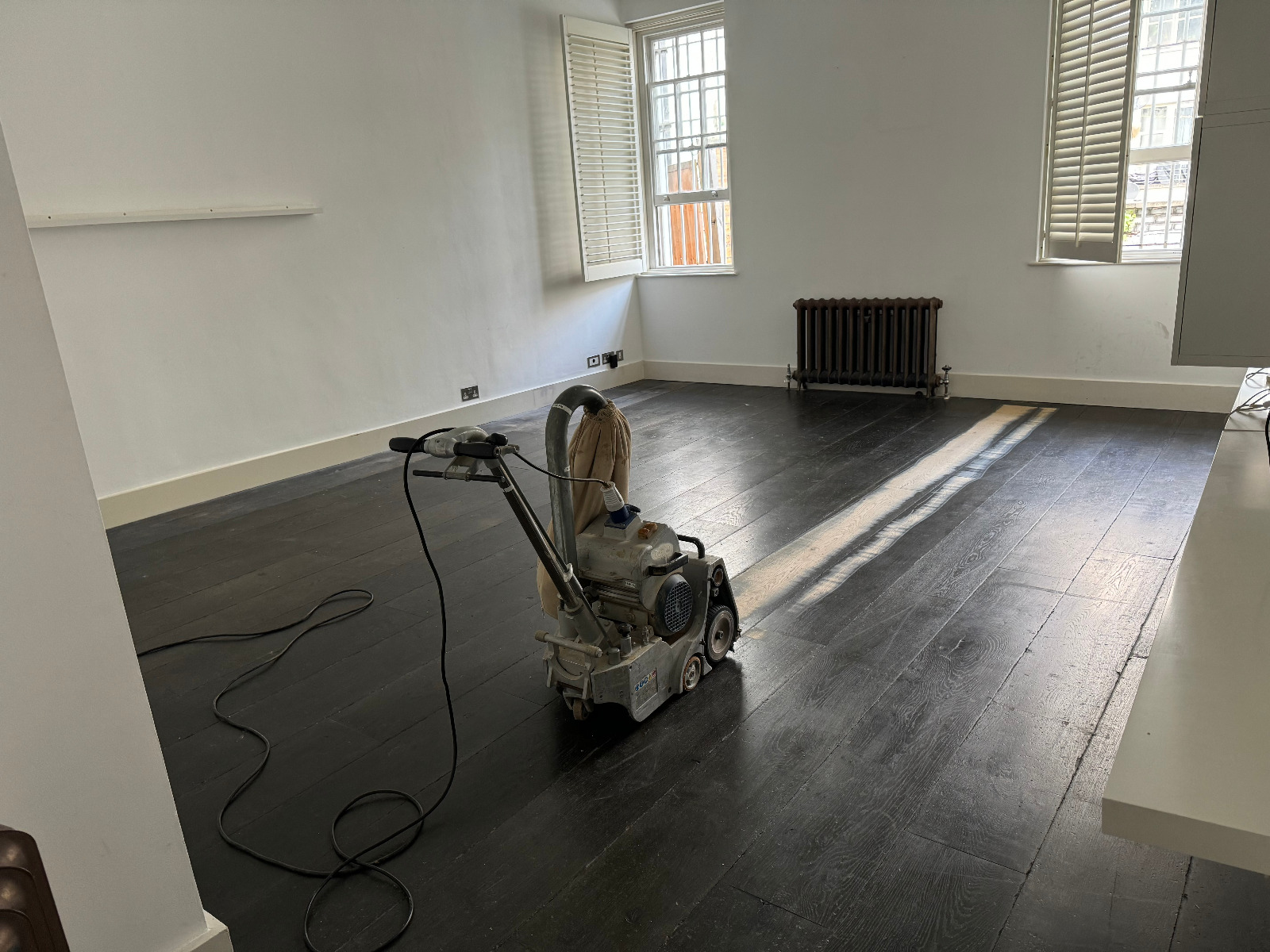 A floor sanding machine is positioned in a living room with dark-stained pine wood flooring, in the process of being refinished. Sunlight streams through the window, highlighting the room's minimalistic decor.