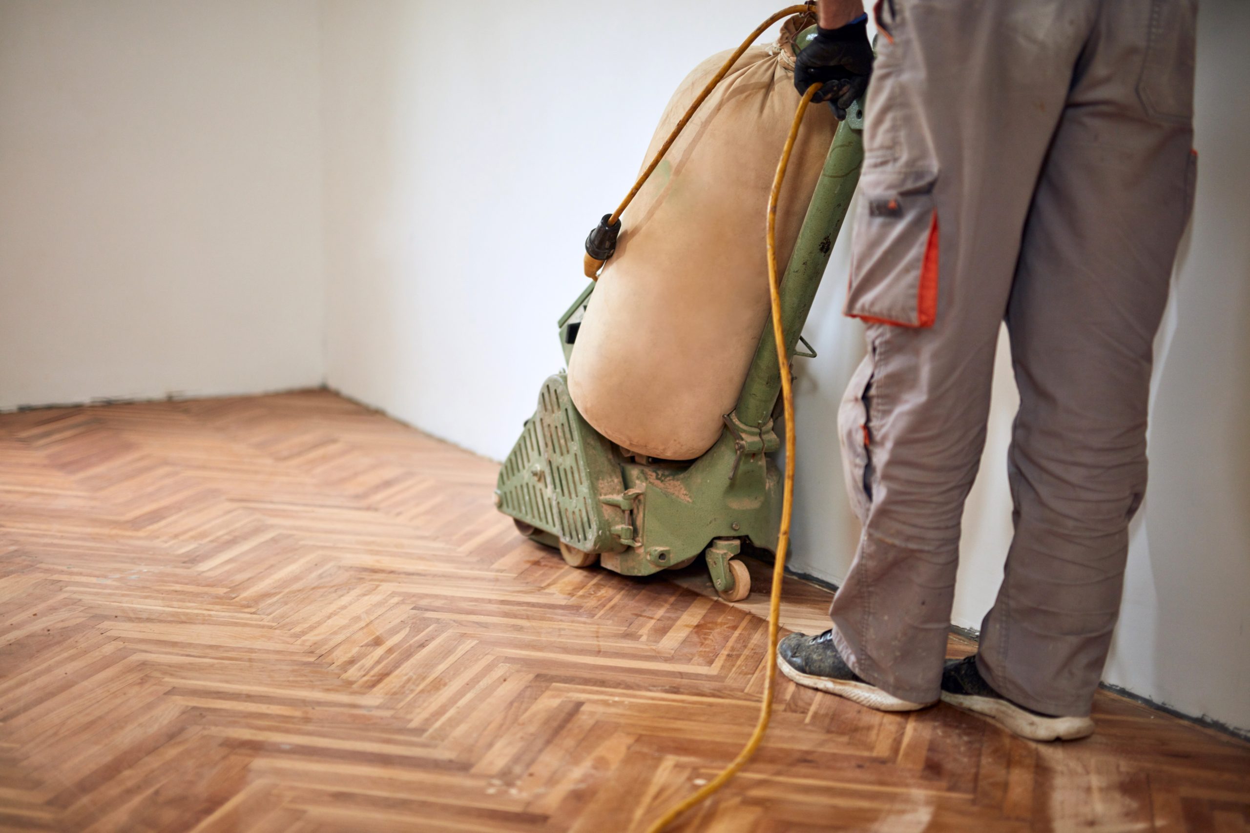 A worker sanding a herringbone parquet wood floor with a drum sander. The floor is in the process of being refinished, with the natural wood tones starting to show after sanding. The person is wearing work pants and sneakers, holding the sander as it operates along the edges of the room.