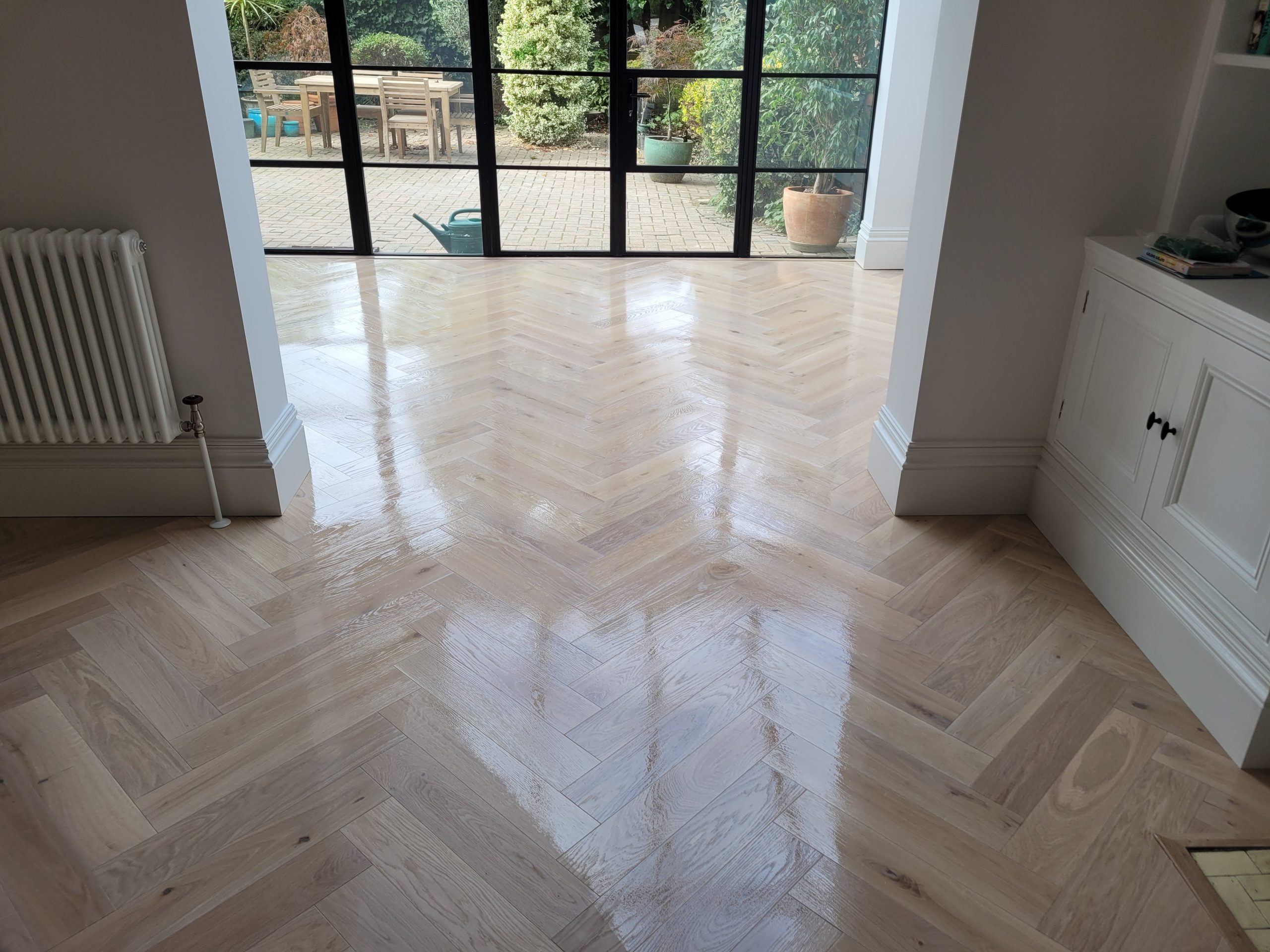 A room with a beautifully polished light herringbone wooden floor that shines under natural light. The floor leads to large glass doors that open to a patio with outdoor furniture and potted plants, creating a seamless transition between indoor and outdoor spaces. The white walls and built-in cabinet add a clean, contemporary feel to the room, while a classic radiator enhances the cozy atmosphere.