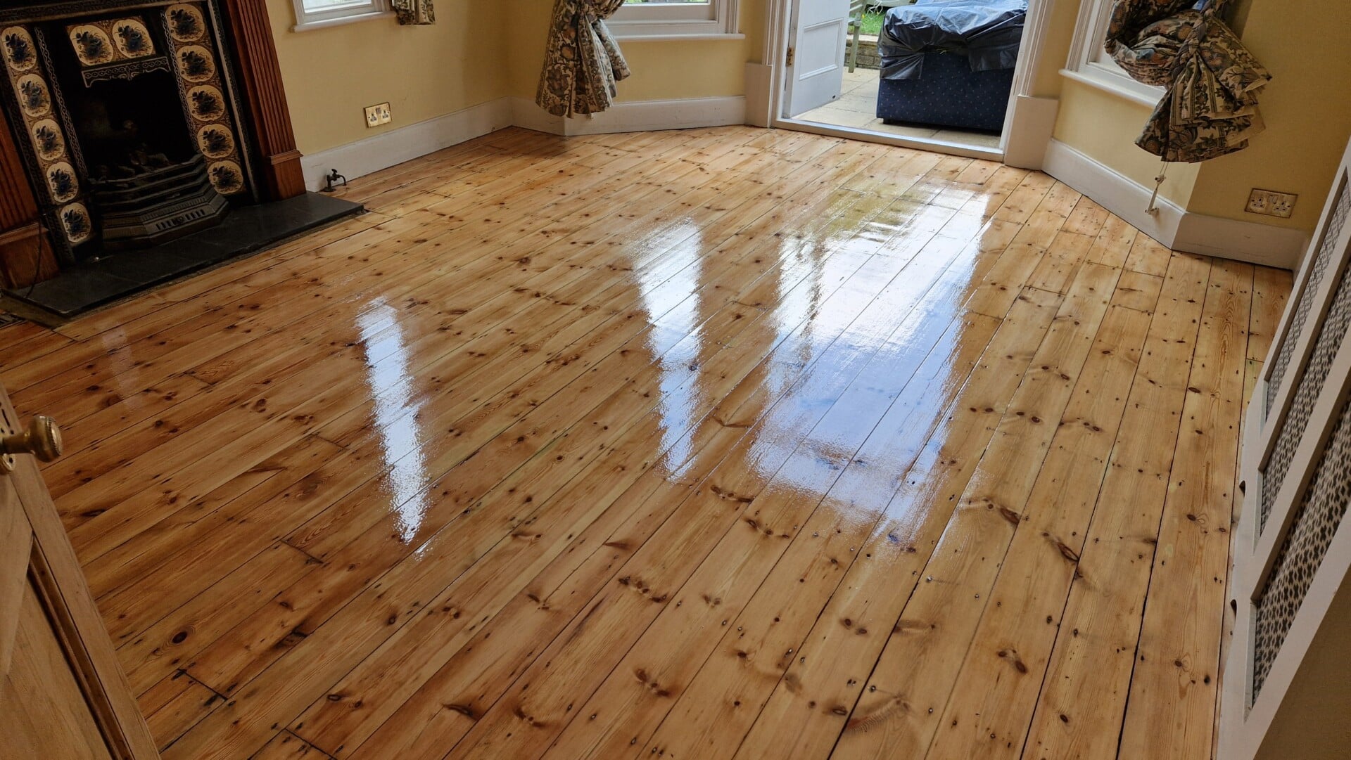 A room with highly polished pine wood flooring, reflecting the natural light streaming in through large windows. The room features a classic fireplace with ornate tiles and a partially open door leading to an outdoor space. The smooth, glossy finish of the floor highlights its freshly sanded and varnished surface.