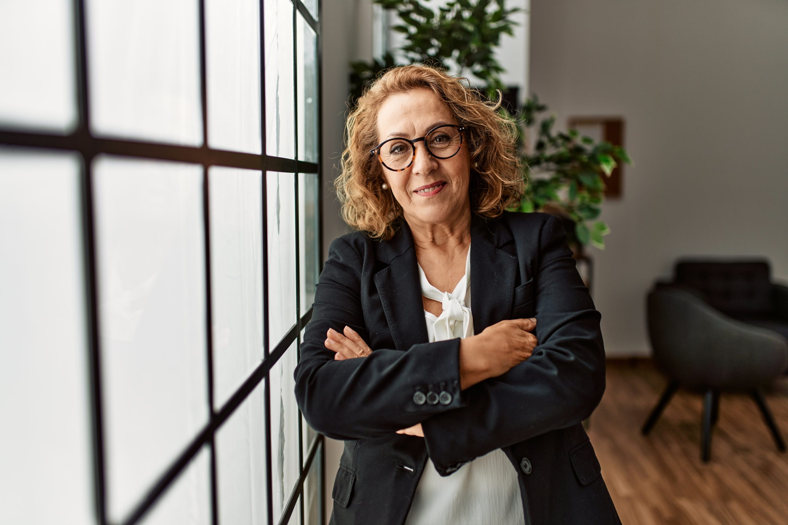 A confident woman stands with her arms crossed, leaning against a large window in a modern office space. The natural light highlights the freshly refinished wooden floor, adding warmth to the sleek and professional environment. Floor sanding and refinishing done by "Sanding Wood Floors" contributes to the clean, polished look of the workspace.