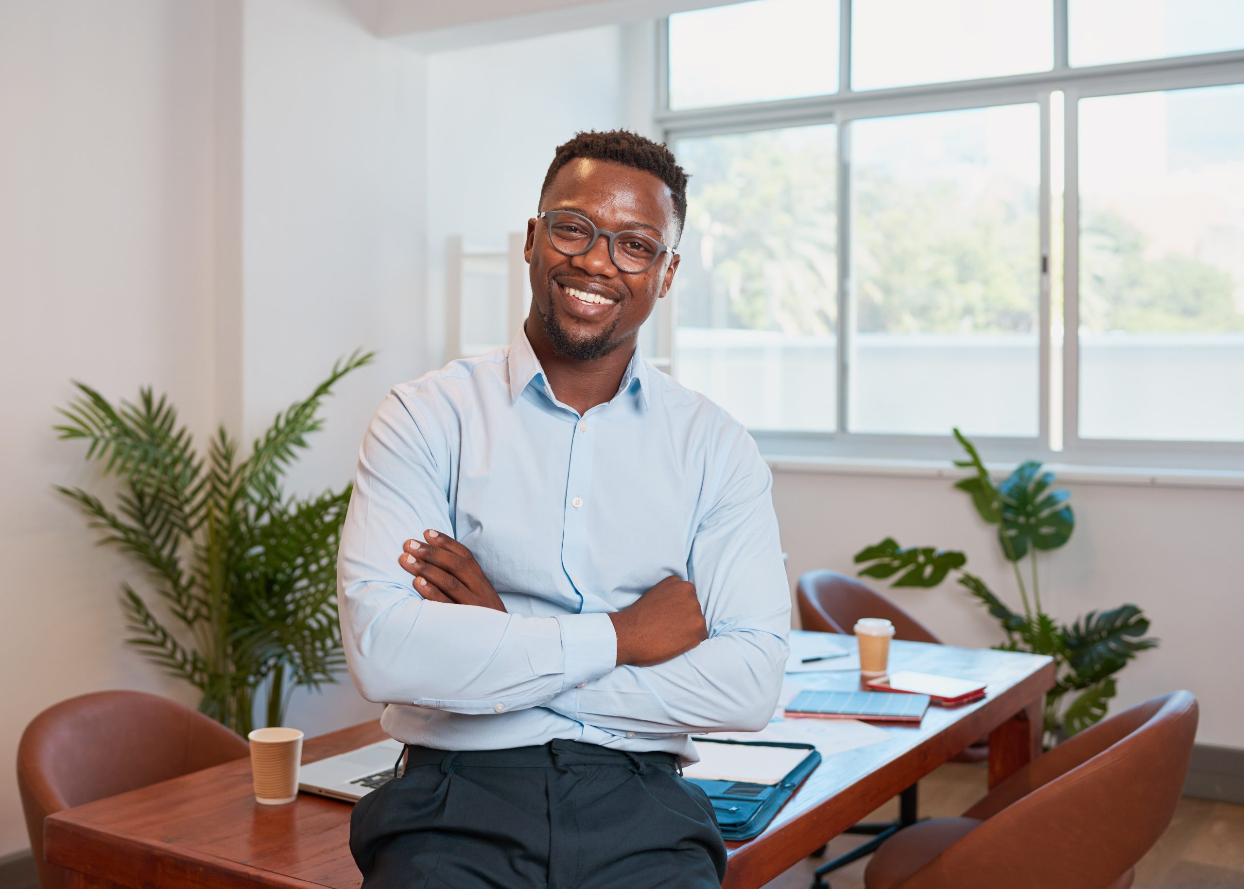 A confident man wearing glasses and a light blue shirt sits on the edge of a wooden desk with his arms crossed, smiling in a bright office space with plants and natural light. The well-sanded and polished wooden floor beneath complements the modern and professional setting, showcasing the high-quality work done by "Sanding Wood Floors."