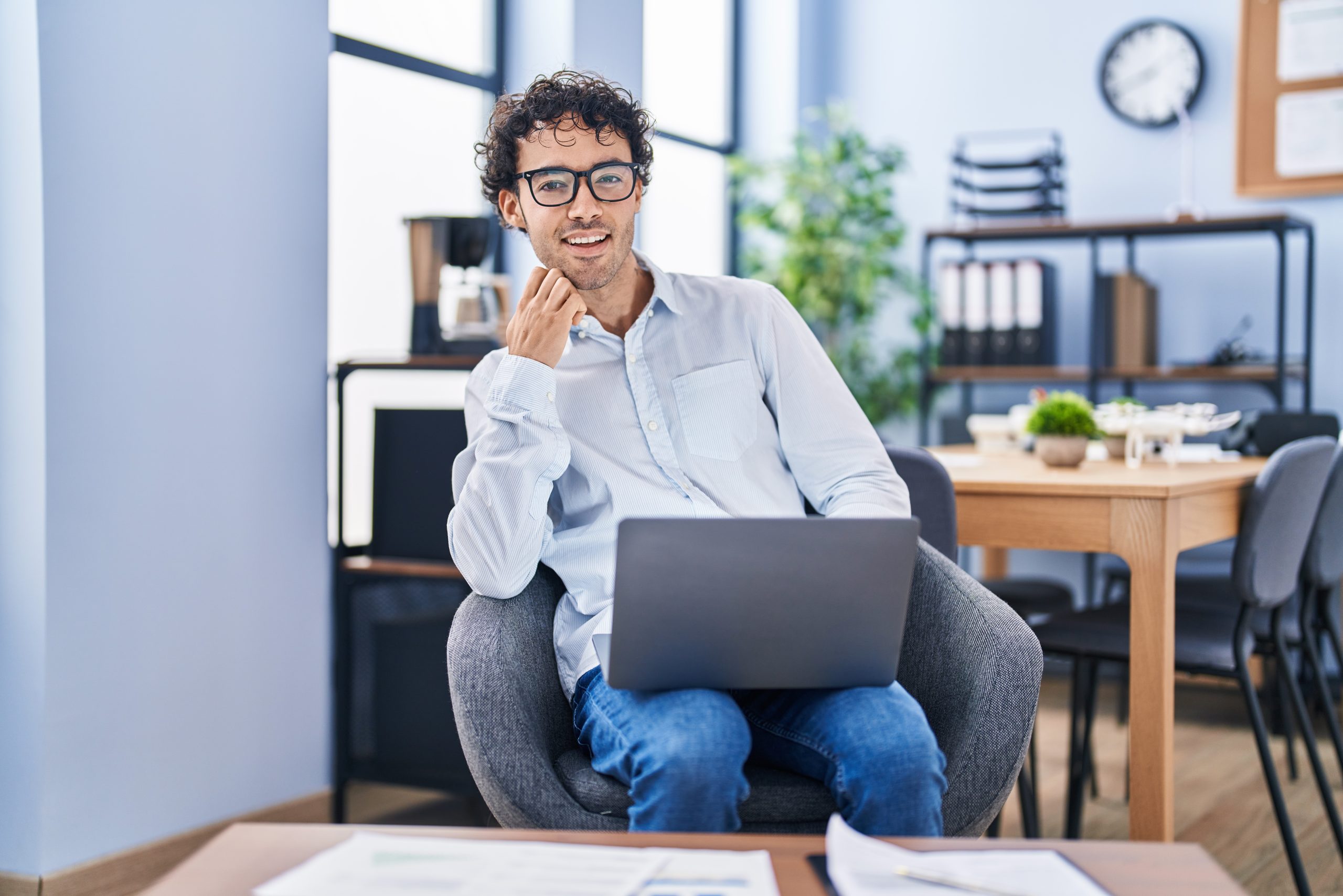 A smiling man with glasses sits comfortably in a modern office chair, working on his laptop. The bright office environment features beautifully refinished hardwood floors that gleam under the soft lighting, demonstrating the expert craftsmanship of floor sanding and refinishing done by "Sanding Wood Floors."
