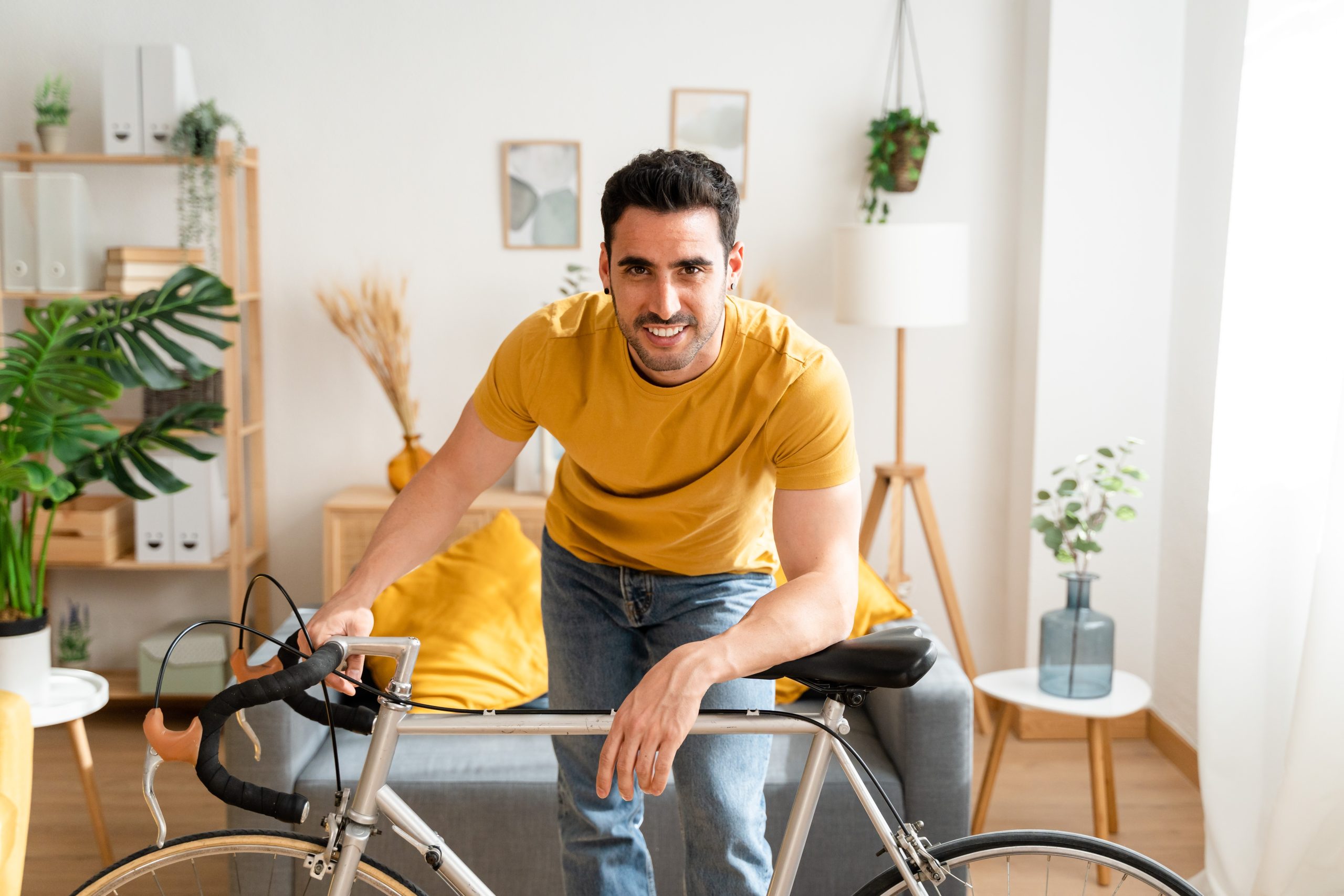 A man in a yellow shirt smiles while leaning over his bicycle in a cozy, modern living room with a beautifully refinished wooden floor. The warm tones of the room and smooth flooring highlight the professional floor sanding and refinishing done by "Sanding Wood Floors."