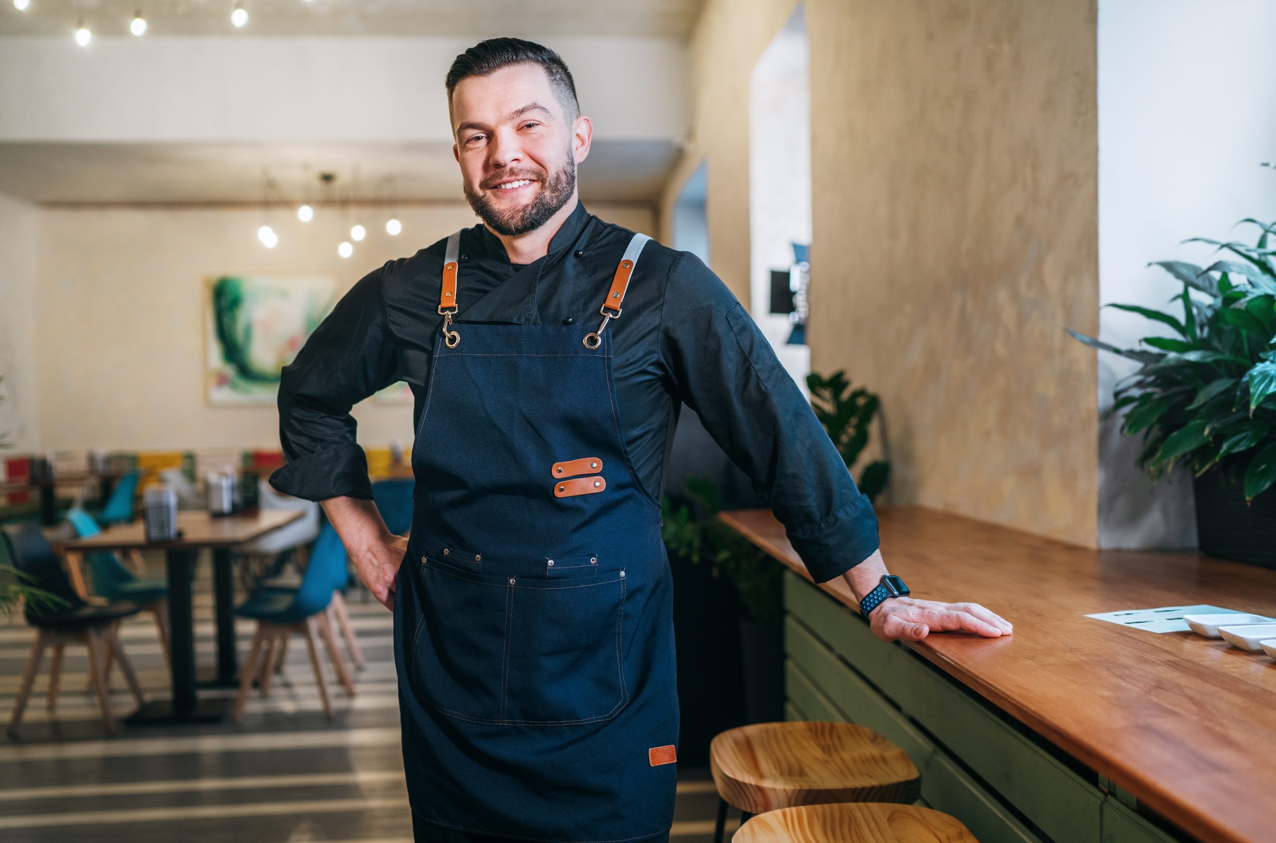 A smiling chef stands confidently in a modern restaurant, wearing a dark apron and resting his hand on a beautifully restored wooden counter. The polished floors and countertops highlight the expert sanding and refinishing work done by "Sanding Wood Floors."