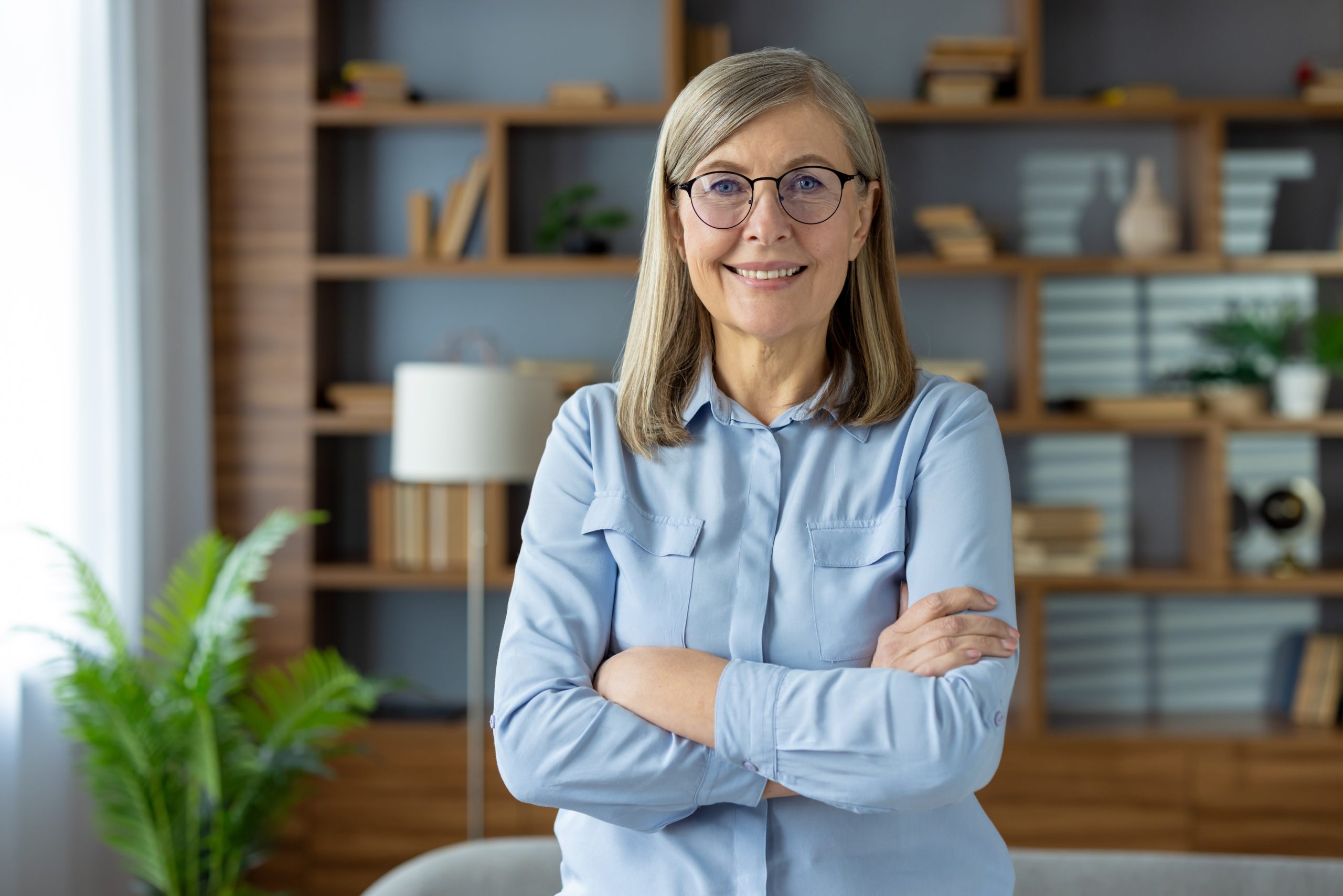 A confident, older woman with glasses stands smiling in a modern, cozy living room. She is wearing a light blue shirt and has her arms crossed. A wooden bookshelf filled with books and plants forms the background, complementing the serene and elegant setting.