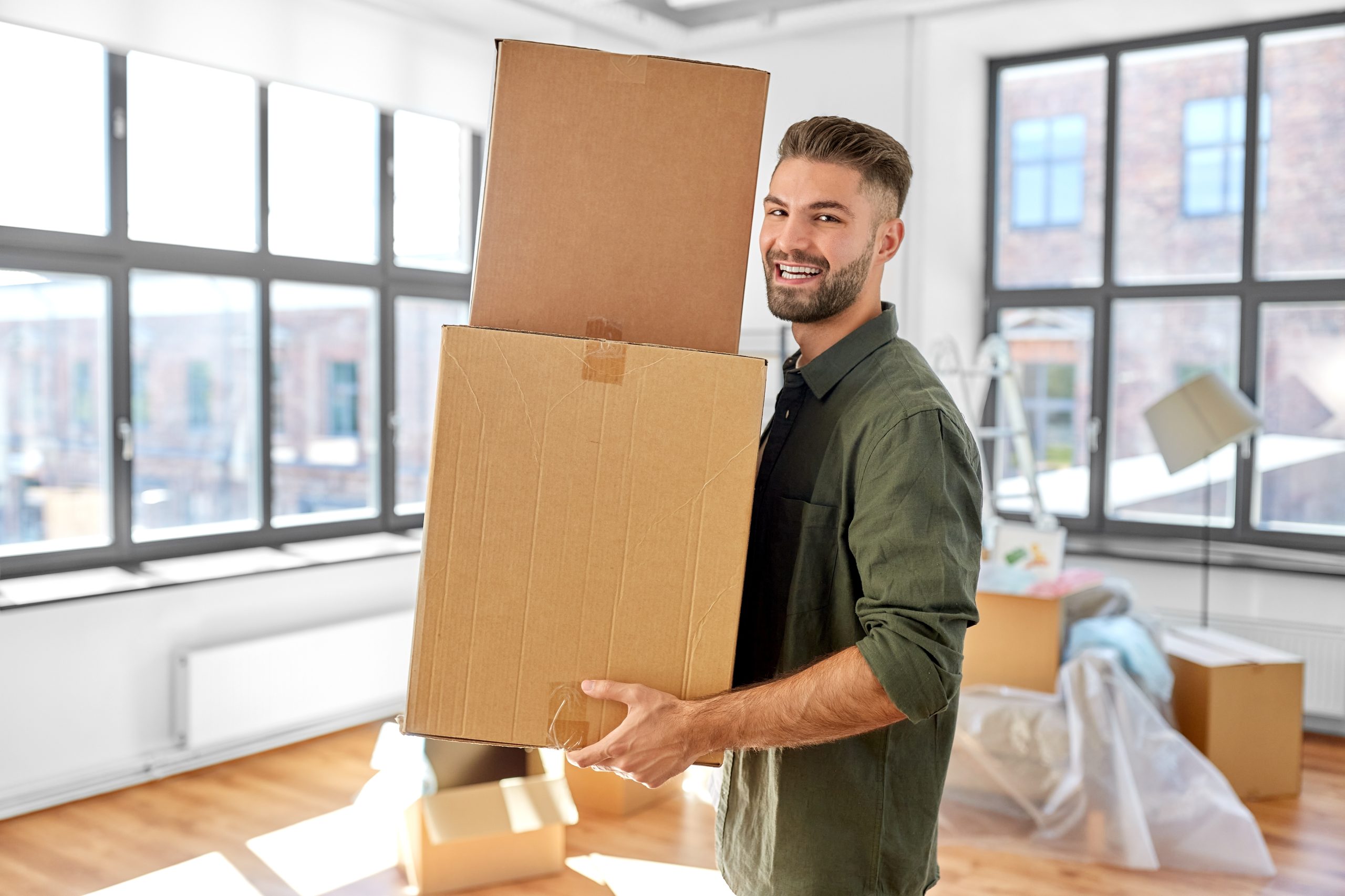 A smiling man carrying two cardboard boxes stands in a bright, modern apartment. The room features large windows, abundant natural light, and beautifully refinished wooden floors with a warm finish.