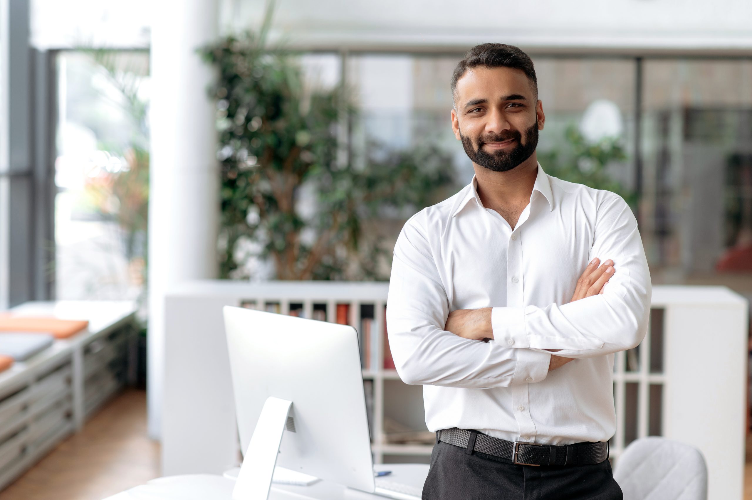 A confident businessman in a white shirt stands with arms crossed in a bright, modern office. The workspace features large windows, lush greenery, sleek furniture, and beautifully refinished wooden floors.