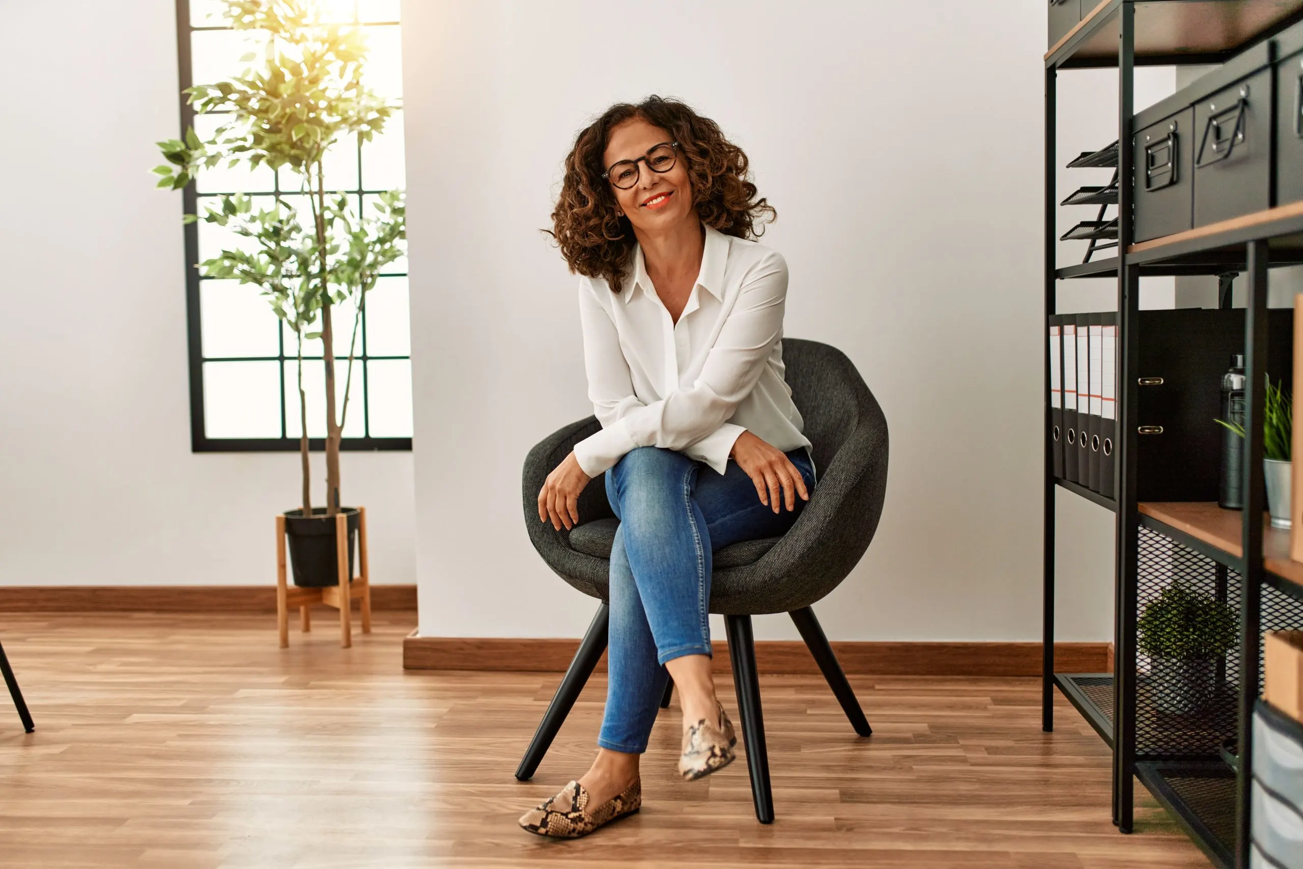 A confident woman with curly hair, wearing glasses and a white blouse, sits on a modern gray chair in a minimalist office space. The room features sleek shelving, a potted plant, and beautifully refinished wooden floors.