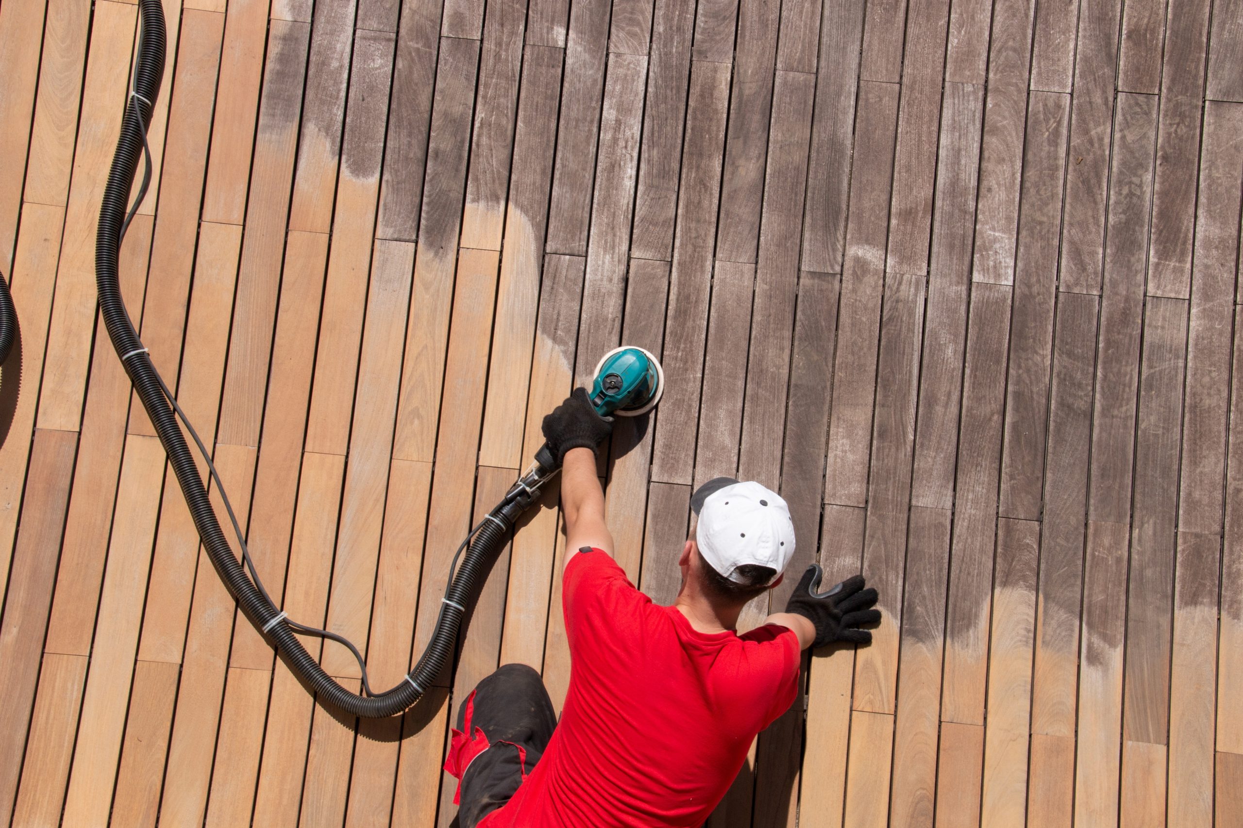 A professional worker refinishing a weathered wooden deck using an electric sander to restore its original luster. The image highlights the precision and effectiveness of floor sanding services provided by "Sanding Wood Floors," transforming dull and worn wood into a refreshed and polished surface.