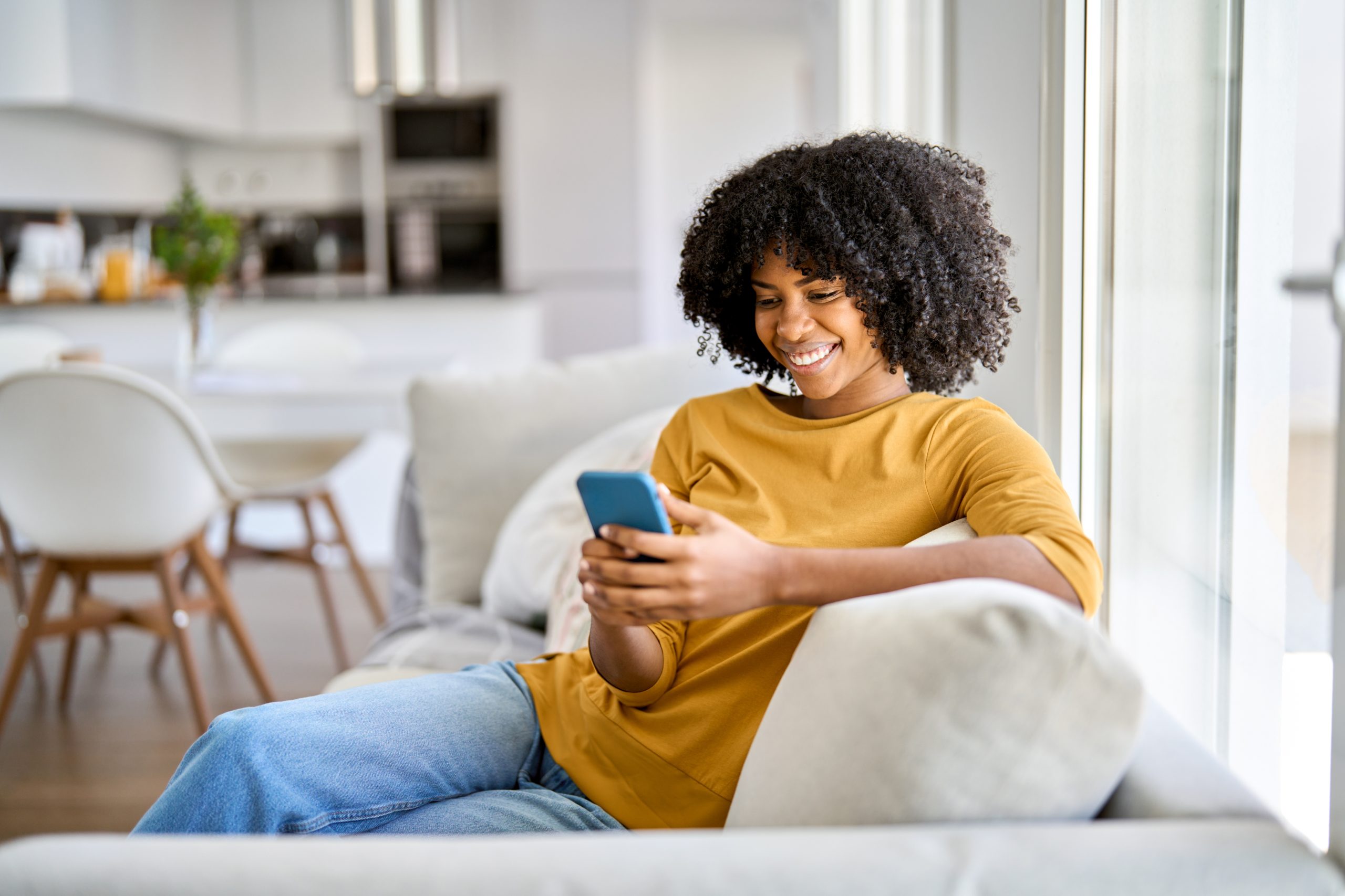 A cheerful woman in a mustard-yellow top relaxes on a cream-colored sofa by a large window, smiling while using her smartphone. The background features a modern dining area and kitchen, with beautifully refinished wooden floors adding warmth to the space.