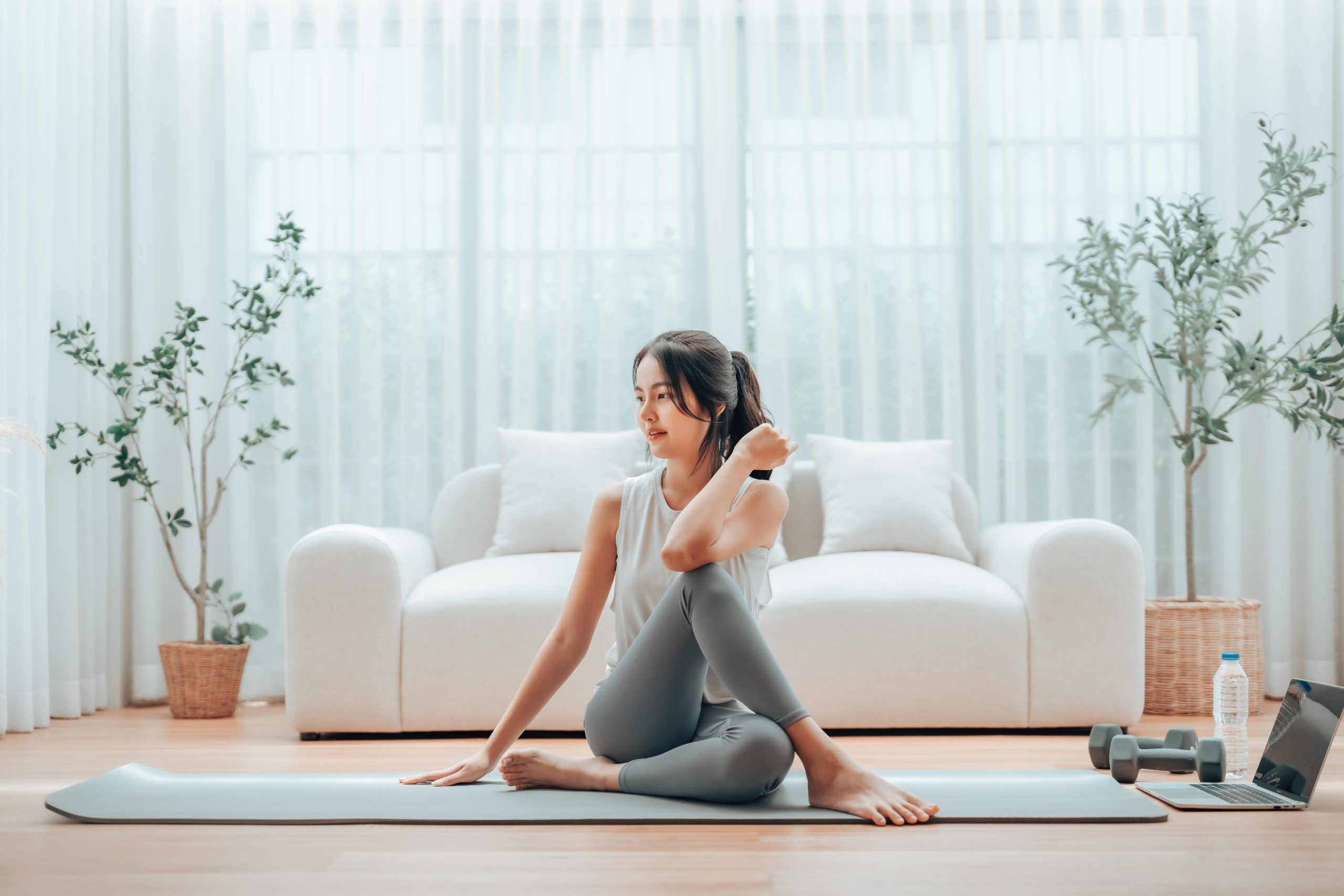 A young woman in workout attire sits on a yoga mat in a bright, airy living room, stretching and practicing yoga. The serene space features a white sofa, potted plants, and beautifully refinished wooden floors.